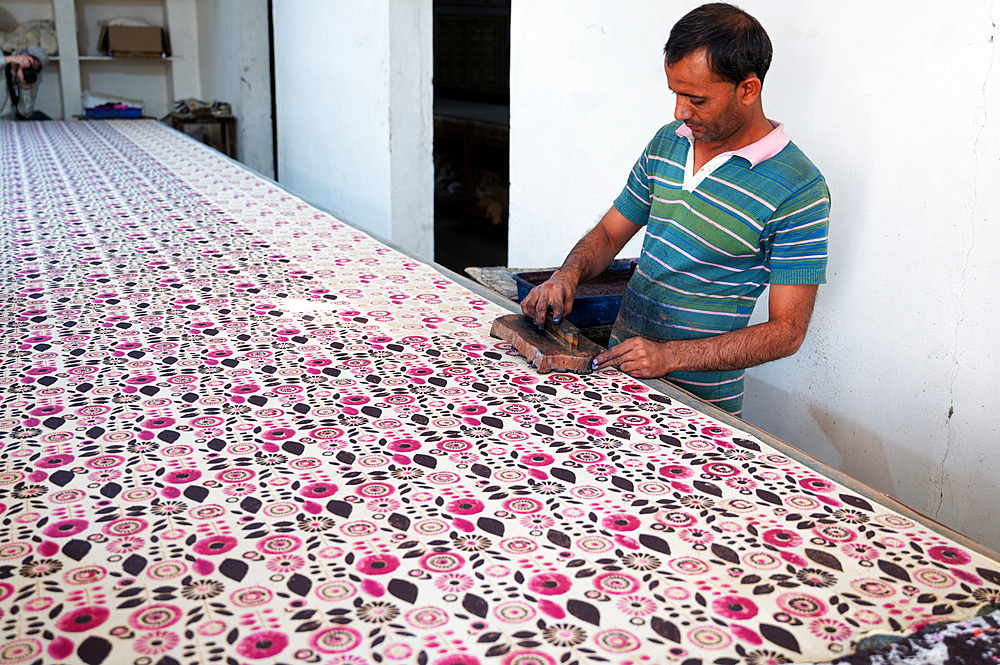 Man using carved and inked wooden blocks to block print large bolt of cotton fabric before rinsing then leaving in the sun to dry.