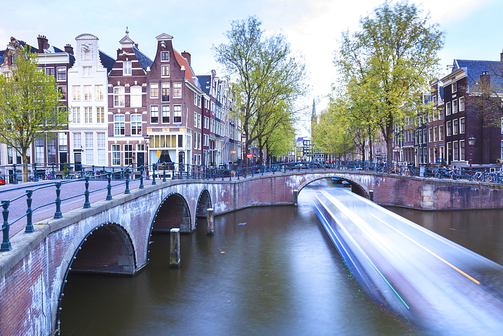 Long exposure of a tourist boat crossing canals Keizersgracht from Leidsegracht at dusk, Amsterdam, Netherlands, Europe 