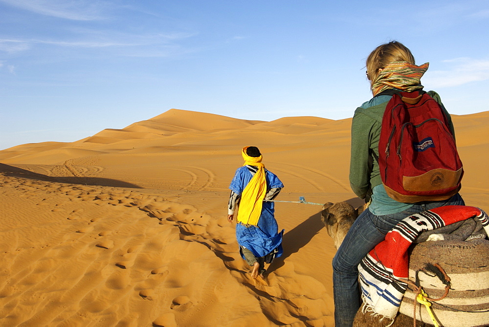 A berber man in traditional outfit leads a woman on a camel through the dunes of Erg Chebbi on the periphery of the Sahara desert in eastern Morocco