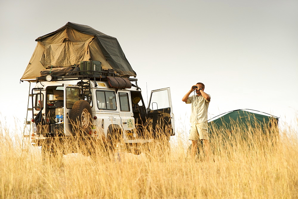 A man looking through binoculars alongside a Land Rover Defender at a campsite in the Kabwoya wildlife reserve in Uganda.