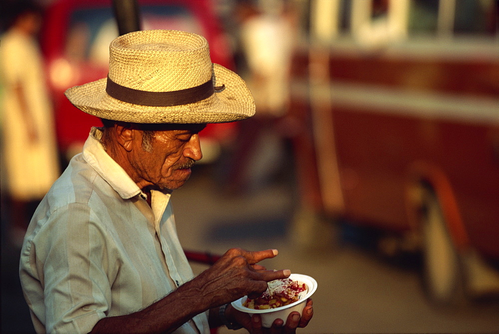 Portrait of an elderly man in a straw hat eating a snack in San Miguel, El Salvador, Central America