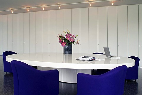 Oval conference table with a laptop and chairs in a modern design in front of a white office wall unit