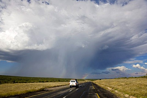 Four-wheel vehicle with a roof tent driving on the Trans Kalahari Highway towards a rainbow, paved road in Namibia, Africa