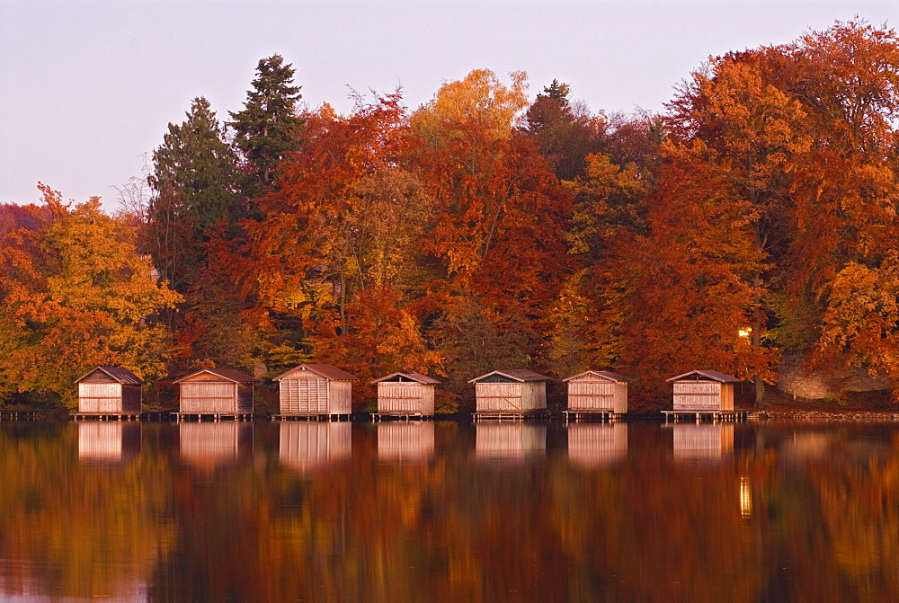 Boathouses and their reflections in the calm waters of lake Wesslinger See surrounded by autumn foliage, Wessling, Bavaria, Germany, Europe