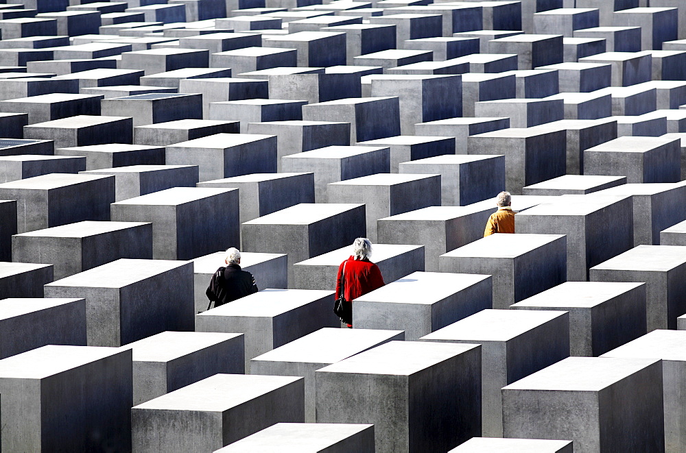 Three people dressed in red, black and yellow-gold jackets walk amongst concrete slabs of the Holocaust Memorial, Berlin, Germany