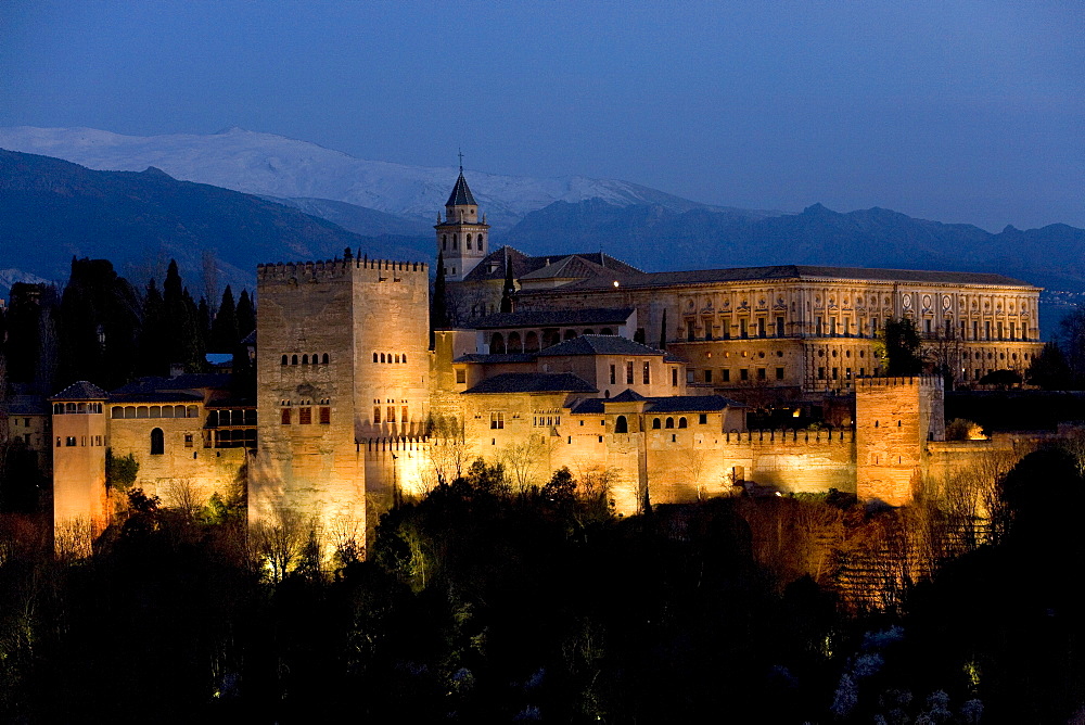 Moorish Alhambra Palace under floodlights at twilight, illuminated at dusk, Sierra Nevada at back, viewed from the Mirador San Nicolas in the El Albayzin or Albaicin quarter of Granada, Andalusia, Spain