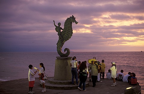 Tourists enjoying the sunset view beneath a statue of a person riding a seahorse on the beach promenade in Puerto Vallarta, Jalisco, Mexico