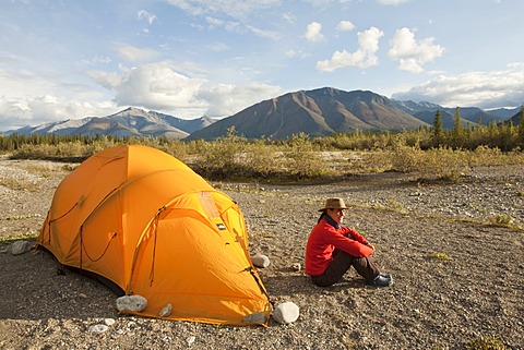 Young woman relaxing, enjoying evening light, sitting beside her tent, Northern Mackenzie Mountains behind, Wind River, Yukon Territory, Canada