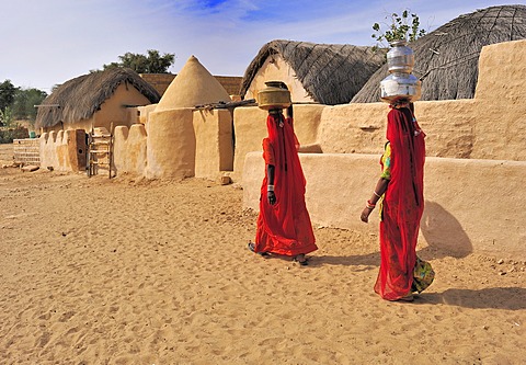 Two Indian women wearing red saris carrying water filled jugs on their heads through a village, Thar Desert, Rajasthan, India, Asia