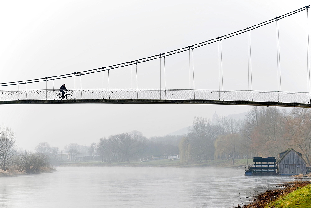 Cyclist crossing a suspension bridge, water mill on the Weser river, Minden, North Rhine-Westphalia, Germany, Europe