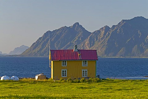 A yellow house in a green meadow near Sanden with steep mountains at back, Lofoten, Nordland, Norway, Europe