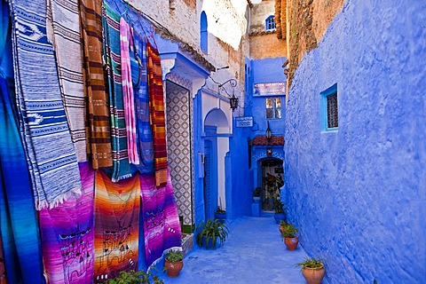 Narrow alleyway in Chefchaouen, walls and footpath are painted in blue, colourful blankets for sale hanging on the wall, Rif Mountains, northern Morocco, Morocco, Africa