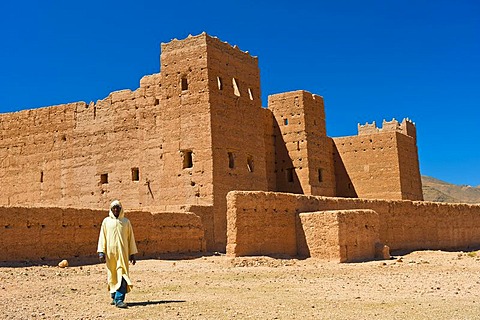 Berber man wearing a traditional djellabah walking in front of Taouirt Kasbah, mud fortress, mud brick building of the Berber tribes, Tighremt, Draa Valley, Southern Morocco, Morocco, Africa