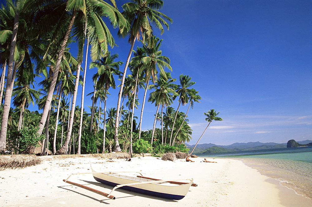 Couple sunbathing on beach with outrigger boat in foreground, El Nido, Bascuit Bay, Palawan, Philippines, Southeast Asia, Asia