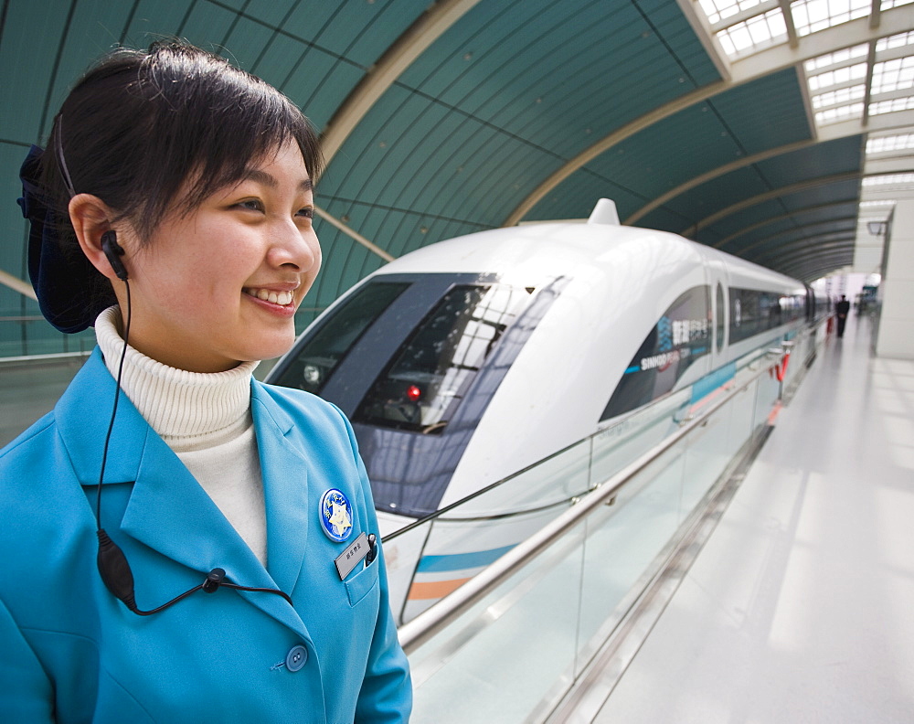 A smiling female attendant wearing a mobile phone earplug standing next to a Magnetic Levitation train on railway platform in Shanghai, China, Asia