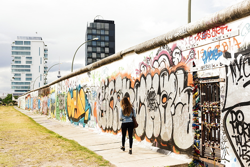 Young woman walking past graffiti on former Berlin Wall, East Side Gallery, Muhlenstrasse, Friedrichshain District, Berlin, Germany, Europe