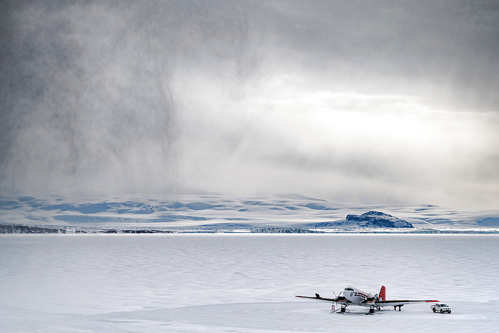 The Basler, stationed on the Terra Nova Bay ice floe in front of the Mario Zucchelli base. The mechanics take advantage of a rare lull to carry out routine maintenance. The aircraft is strapped to the ground. A squall arrives from the left. Victoria Land, Antarctica