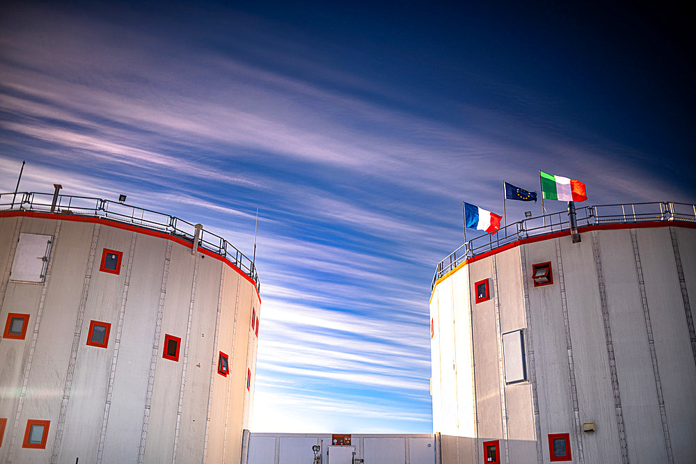 The two towers of the Concordia station, with the French, Italian and European flags flying in the wind, under a blue sky veiled with cirrus clouds aligned in the direction of the wind. The two towers are linked by a tunnel. They are permanently heated to 20°C by the diesel engine that generates the station's electricity. The left tower is the "quiet tower", housing the hospital, bedrooms;34 beds) and offices. The right tower is the "noisy tower": it houses a small workshop, the emergency generator, the waste room, the technical office, the video room, the living room, the kitchens and the refectory. The two towers are about ten meters apart to prevent the risk of fire spreading. Fire is an Antarctic expeditionary's worst nightmare; the French have a very bad experience of it. Concordia Antarctic Research Station, Dome C plateau, East Antarctica.