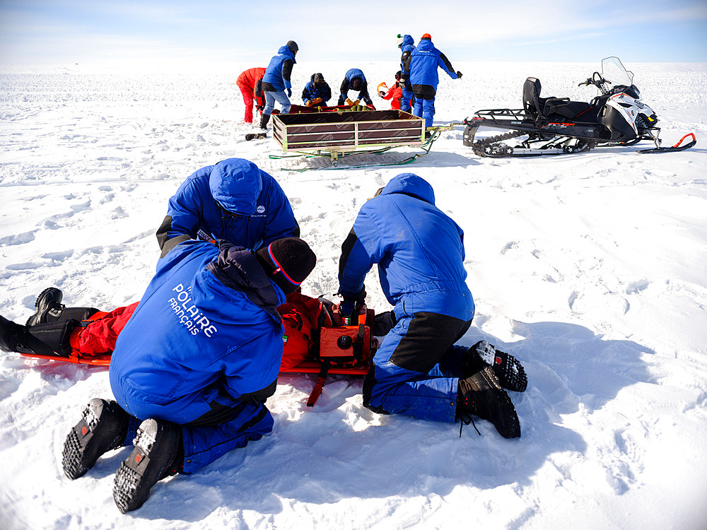 Concordia staff on their knees helping a person lying on a stretcher. Helped by a snowmobile. Rescue exercise in the event of an accident involving several victims. These frequent exercises are supervised by the doctor. All the resort's winter residents are assigned a very specific role in the event of an accident, illness or fire. A redundancy system allows this system to function in the event of one or more winterers failing to respond. Concordia Antarctic Research Station, Dome C plateau, East Antarctica.