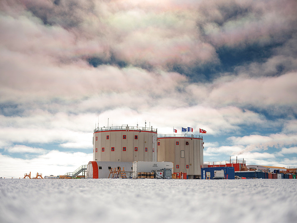 Concordia Station, Italian, European, French, Swiss and German flags. Tent, fire escape, containers, kerosene tanks, smoke from the power station, iridescent clouds. The station under a sky lightly covered with low, iridescent clouds. Concordia Antarctic Research Station, Dome C plateau, East Antarctica.