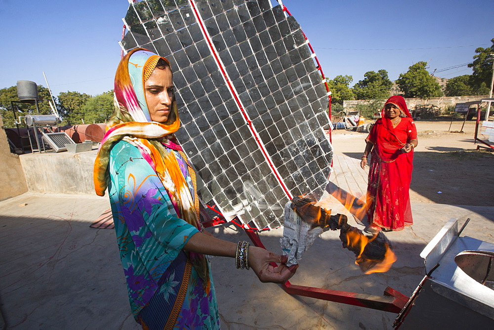 Women constructing solar cookers at the Barefoot College in Tilonia, Rajasthan, India, demonstrate how hot the device is, by holding a sheet of newspaper which instantly sets on fire in the 300 degree Celcius heat. The Barefoot College is a worldwide charity, founded by Bunker Roy, its aims are, education, drinking water, electrification through solar power, skill development, health, women empowerment and the upliftment of rural people. The use of the cookers, vastly reduces the amount of fire wood women have to go out and collect from the forest.