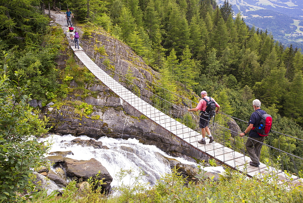 Walkers on the Tour Du Mont Blanc cross a suspension bridge across the meltwater river from the Bionnassay glacier;