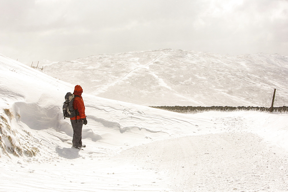 A walker stands next to massive snow drifts blocking the Kirkstone Pass road above Ambleside in the Lake District, Cumbria, England, United Kingdom, Europe