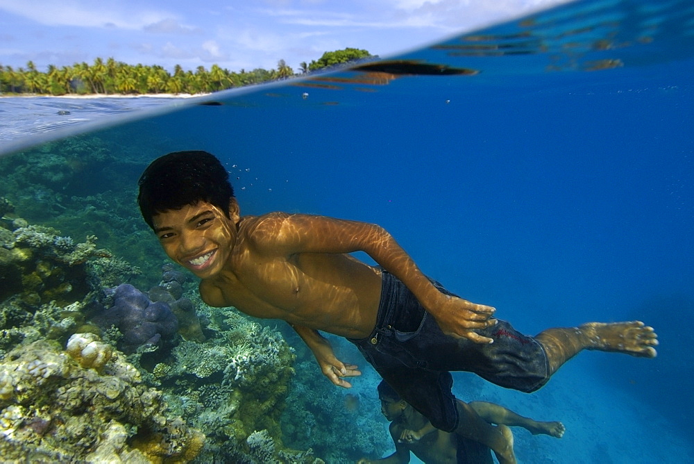 Marshallese boy smiling underwater next to coral reef and coconut trees lining the shore of  Majikin Island, Namu atoll, Marshall Islands, Micronesia, Pacific