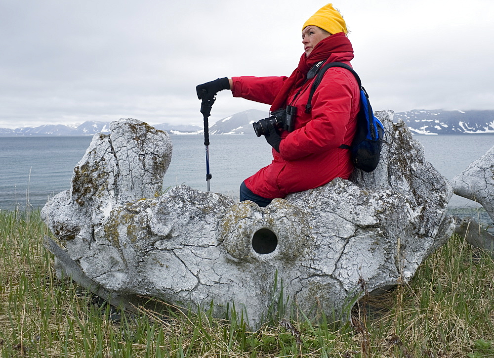 Whalebone Alley, female tourist sitting on bowhead whale bone, Itygran Island (Bering Sea) Russia, Asia.  MORE INFO: Whale Bone Alley was discovered by Soviet archaeologists in 1976, but has remained untouched since and little is known of this place. There is a long double line of bowhead whale bones -- jaws and ribs -- running parallel along the shore for hundreds of yards. Many of the bones, especially the enormous jaw bones, are still standing, propped up by lichen-covered rocks. The location is thought to have been used in about 1300 as a ceremonial site, for a men's secret society or feasting site.