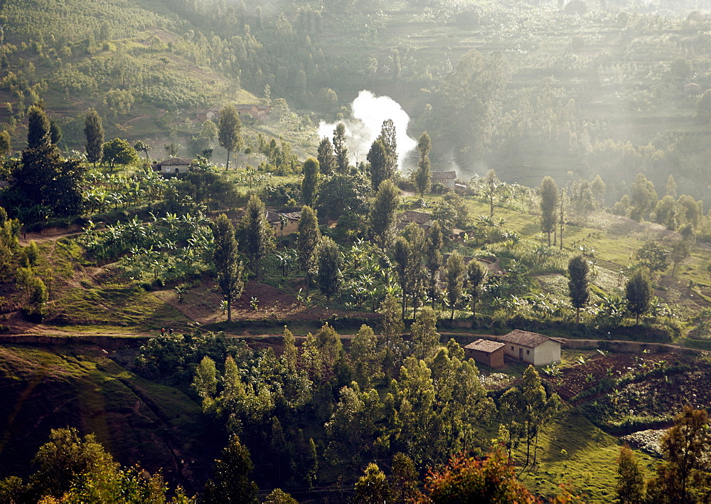 A fire is smoking in the glow of the low sunlight.  The small manageable fields are like a patchwork quilt with the colours of the different crops laid out over "the land of 1000 hills". Gikongoro, Rwanda, East Africa