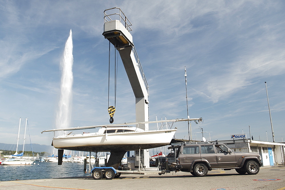 Sailing boat preparing to be lowered into the water by a crane.  Jet d'Eau in the background, Lake Geneva. Geneva, Switzerland