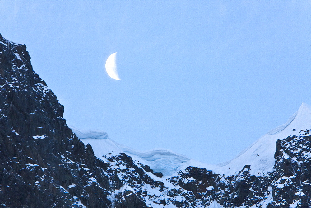 A waxing moon showing over snow covered cliffs in the Lemaire Channel on the west side of the Antarctic peninsula.