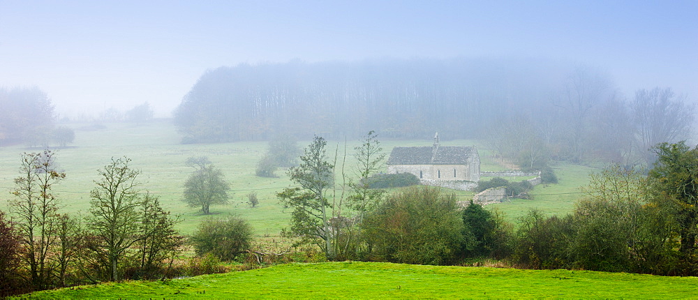 Misty scene and St Oswald's Church, a remote chapel in a field, at Widford near Burford in The Cotswolds, Oxfordshire, UK