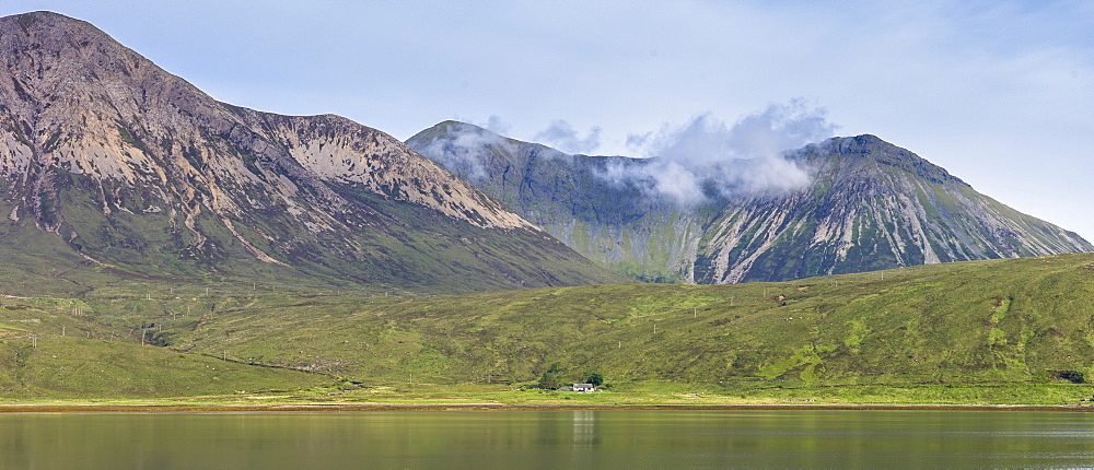 Small white solitary crofters cottage nestling below mountain range reflected in waters of the loch, Isle of Skye, Inner Hebrides, Highlands and Islands, Scotland, United Kingdom, Europe