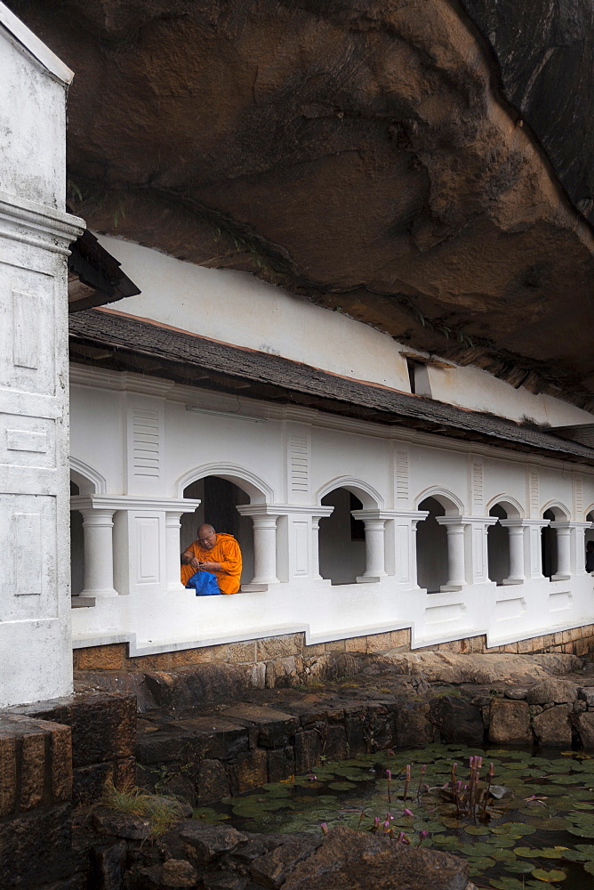 A monk sits and looks at his phone beneath shelter, Royal Rock Temple, Golden Temple of Dambulla, UNESCO World Heritage Site, Dambulla, Sri Lanka, Asia 