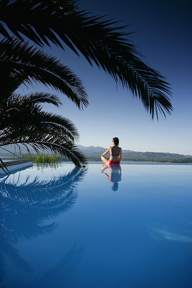 Woman sitting at edge of outdoor pool, sea in back, Bay of Porto Vecchio, Southern Corse, France