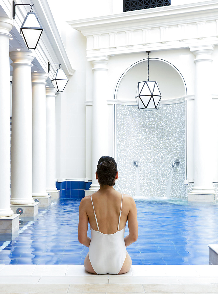 Woman in white bathing suit, sitting with her back to the camera, on the edge of an interior pool. Bath, United Kingdom