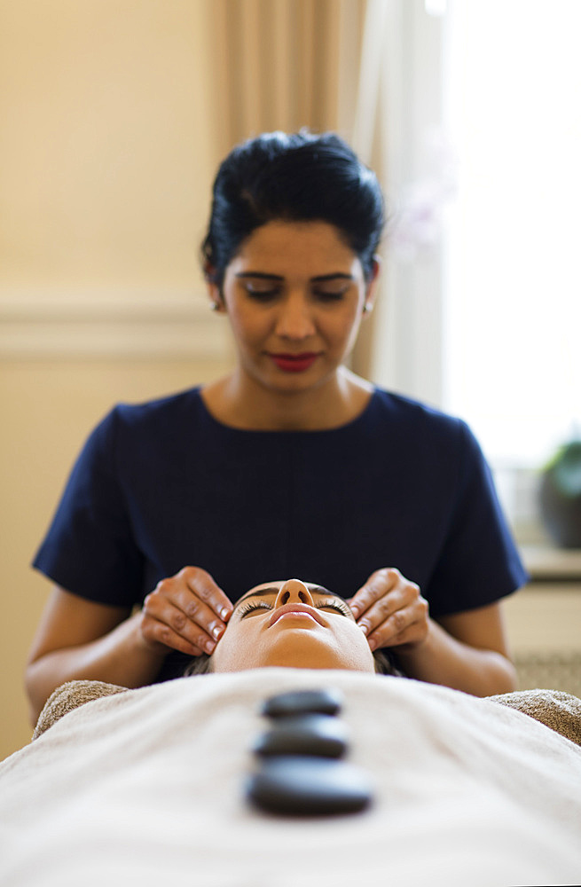 A spa therapist performing a cranial massage, while guest enjoys hot stones on her body. Bath, United Kingdom