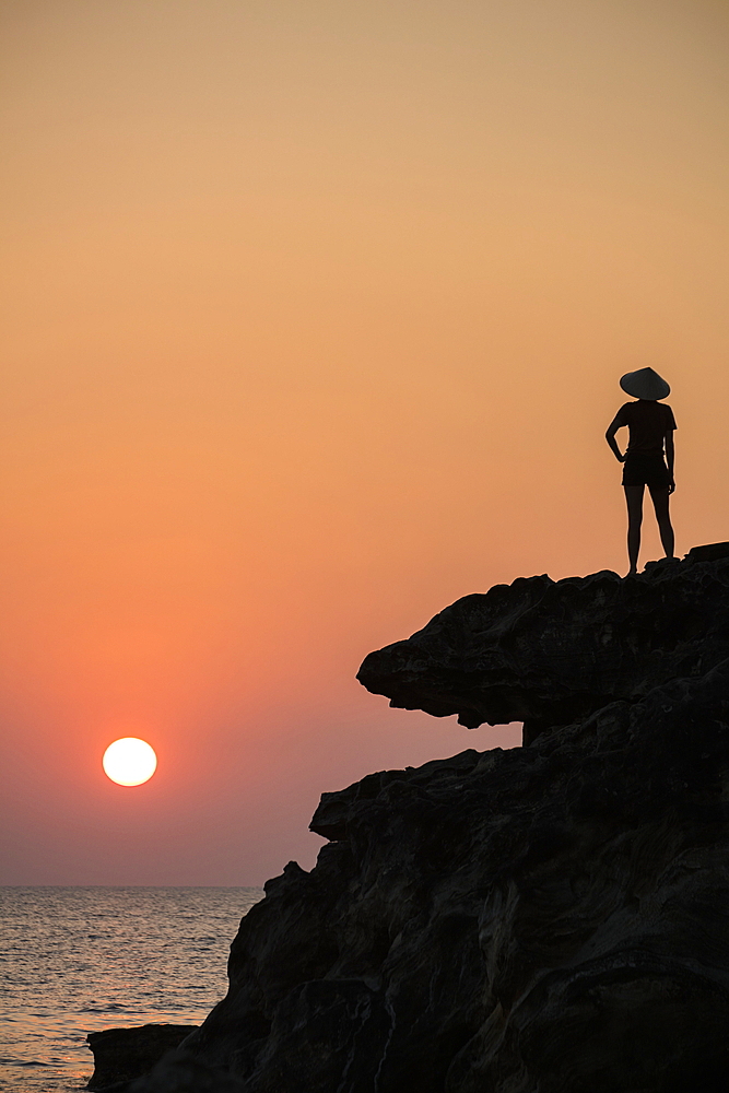 Silhouette of young woman wearing conical hat and looking out to sea from rock ledge next to Dinh Cao Shrine at sunset, Duong Dong, Phu Quoc Island, Kien Giang, Vietnam, Asia