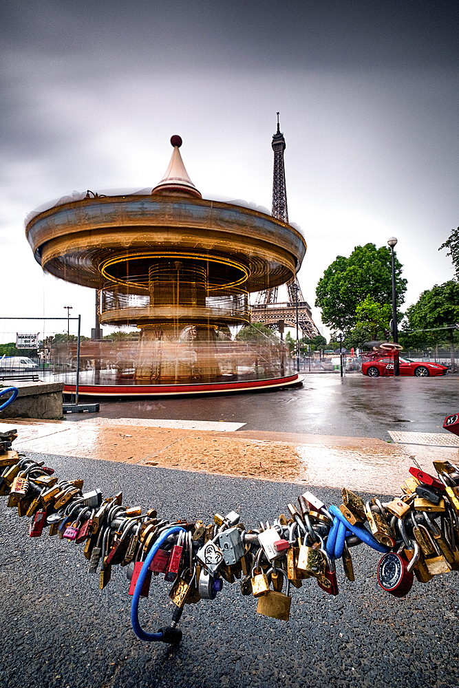 Carousel in front of the Eiffel Tower in rainy weather; in the foreground, love-locks are fastened to the chain of street-bluffing; Paris; Île-de-france; France