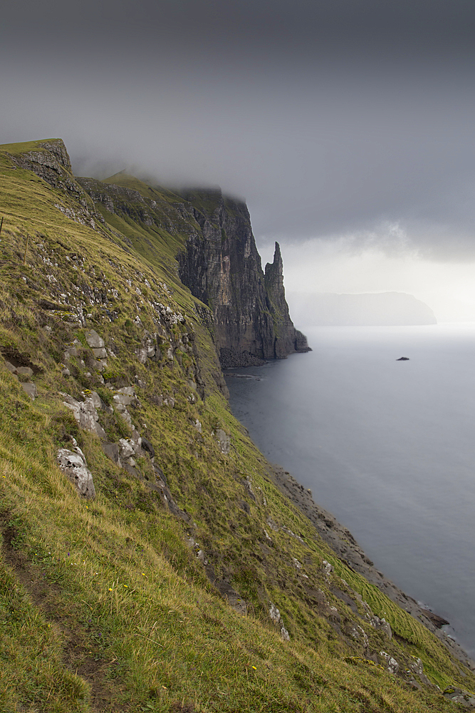 View along cliffs to Witches Finger, sea stack, Vagar, Faeroer, dark rain clouds.