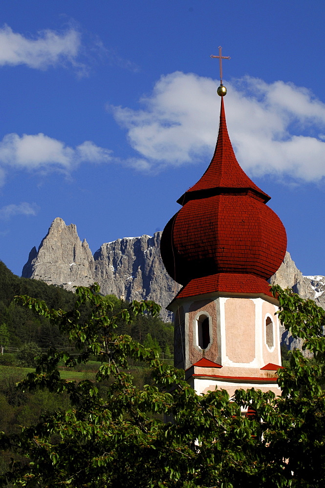 The steeple of Saint Oswald church between green branches and mountains, Kastelruth, South Tyrol, Italy, Europe