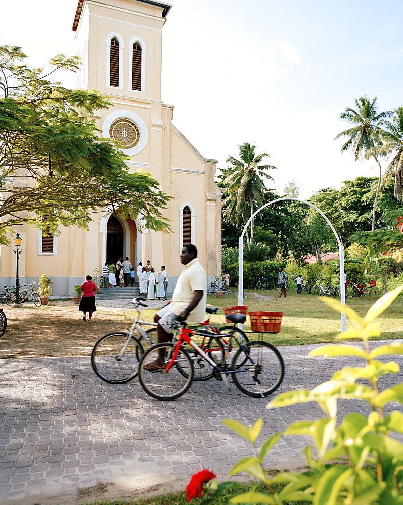 Man with rental bikes passing in front of the church at La Passe, La Digue, La Digue and Inner Islands, Republic of Seychelles, Indian Ocean