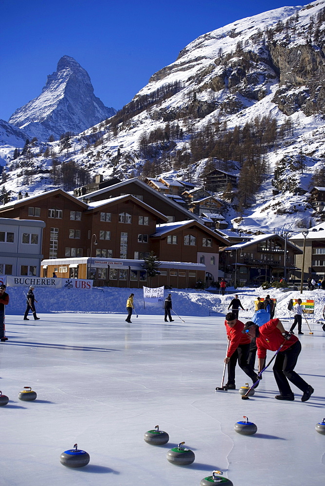 People curling on a rink, Matterhorn in background, Zermatt, Valais, Switzerland (Curling: A rink game where round stones are propelled by hand on ice towards a tee (target) in the middle of a house (circle)).