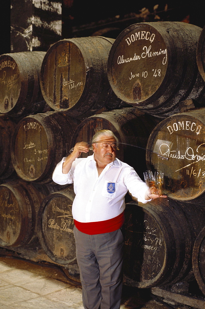 A wine taster pouring samples of Sherry into tasting glasses in front of a heap of Sherry casks, in the Bodegas Pedro Domeq, Jerez de la Frontera, Cadiz province, Andalusia, Spain