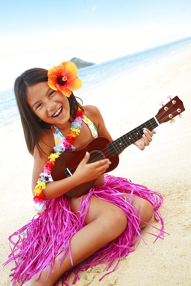 Hawaii, Oahu, Young girl smiling and playing ukulele on the beach in a hula skirt.