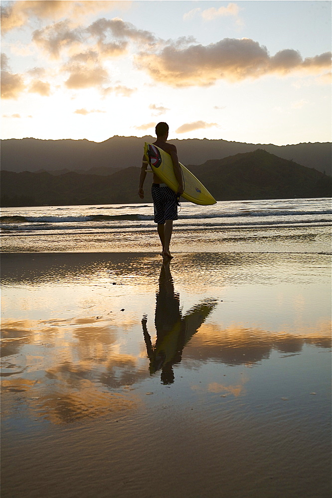 Hawaii, Kauai, Hanalei Bay, young man at the beach walking with surfboard at sunset.