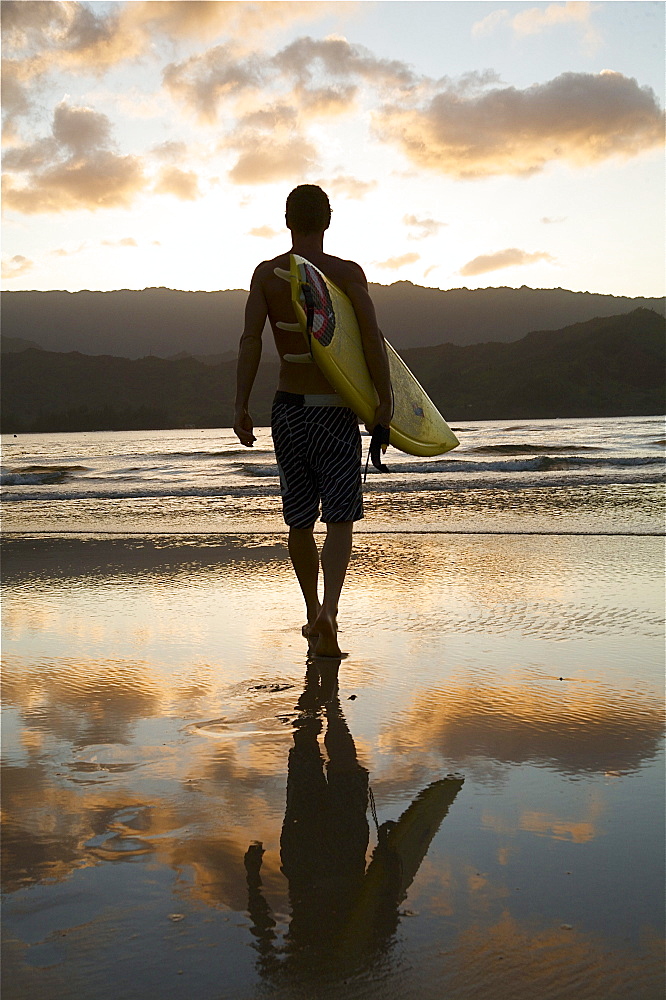 Hawaii, Kauai, Hanalei Bay, young man at the beach walking with surfboard at sunset.