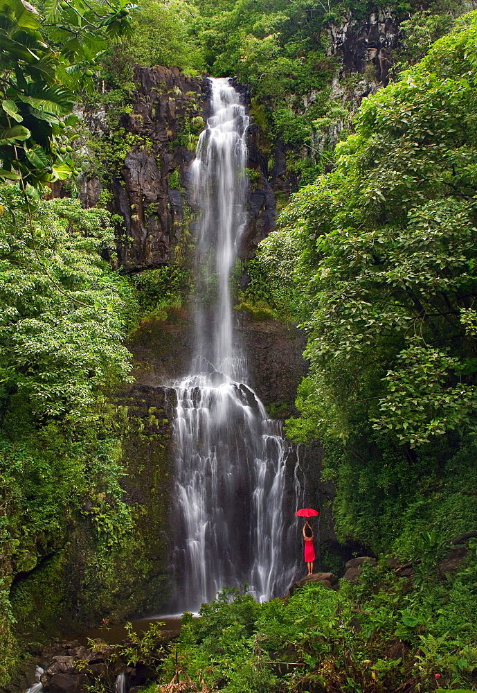 Hawaii, Maui, Kipahulu, Hana Coast, Woman stands with umbrella at Wailua Falls surrounded by foliage.