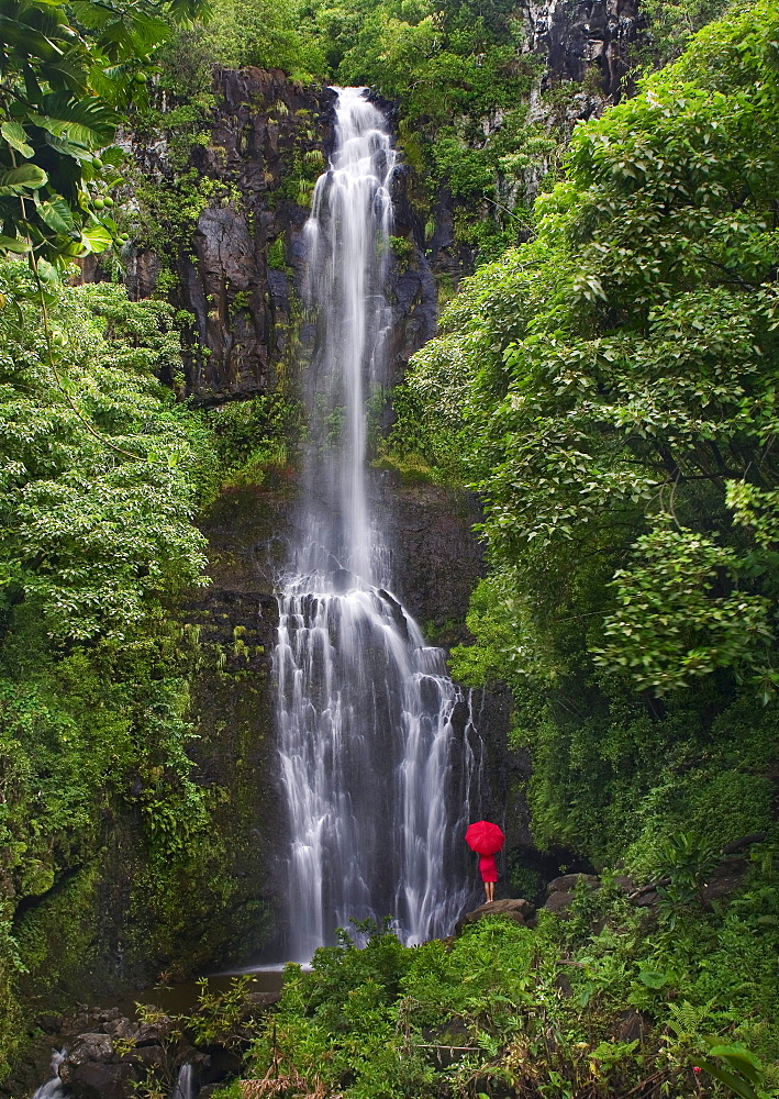 Hawaii, Maui, Kipahulu, Hana Coast, Woman stands with umbrella at Wailua Falls surrounded by foliage.